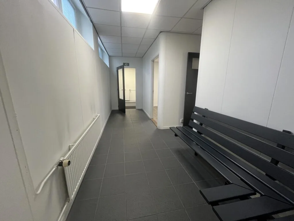 Gang met zwarte tegelvloer, witte muren, een zwarte bank en openstaande deuren in een gebouw aan de Burgemeester Smitsstraat.